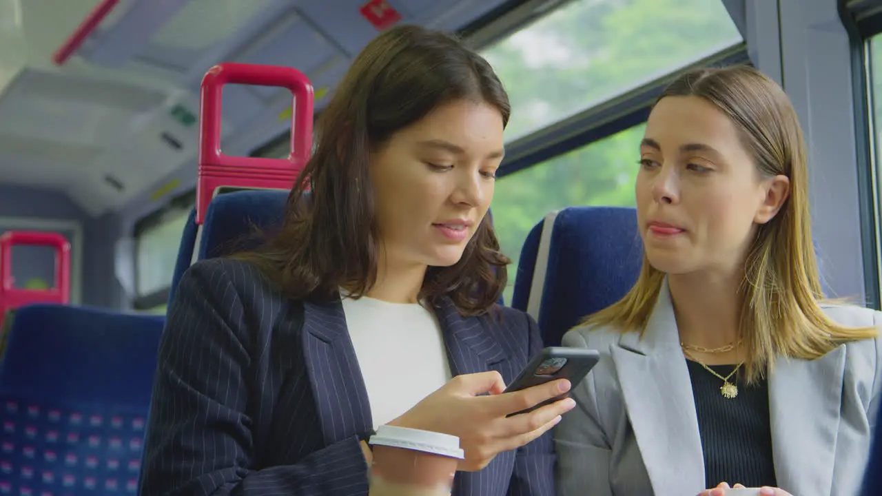 Two Businesswomen With Takeaway Coffees Commuting To Work On Train Looking At Mobile Phone Together