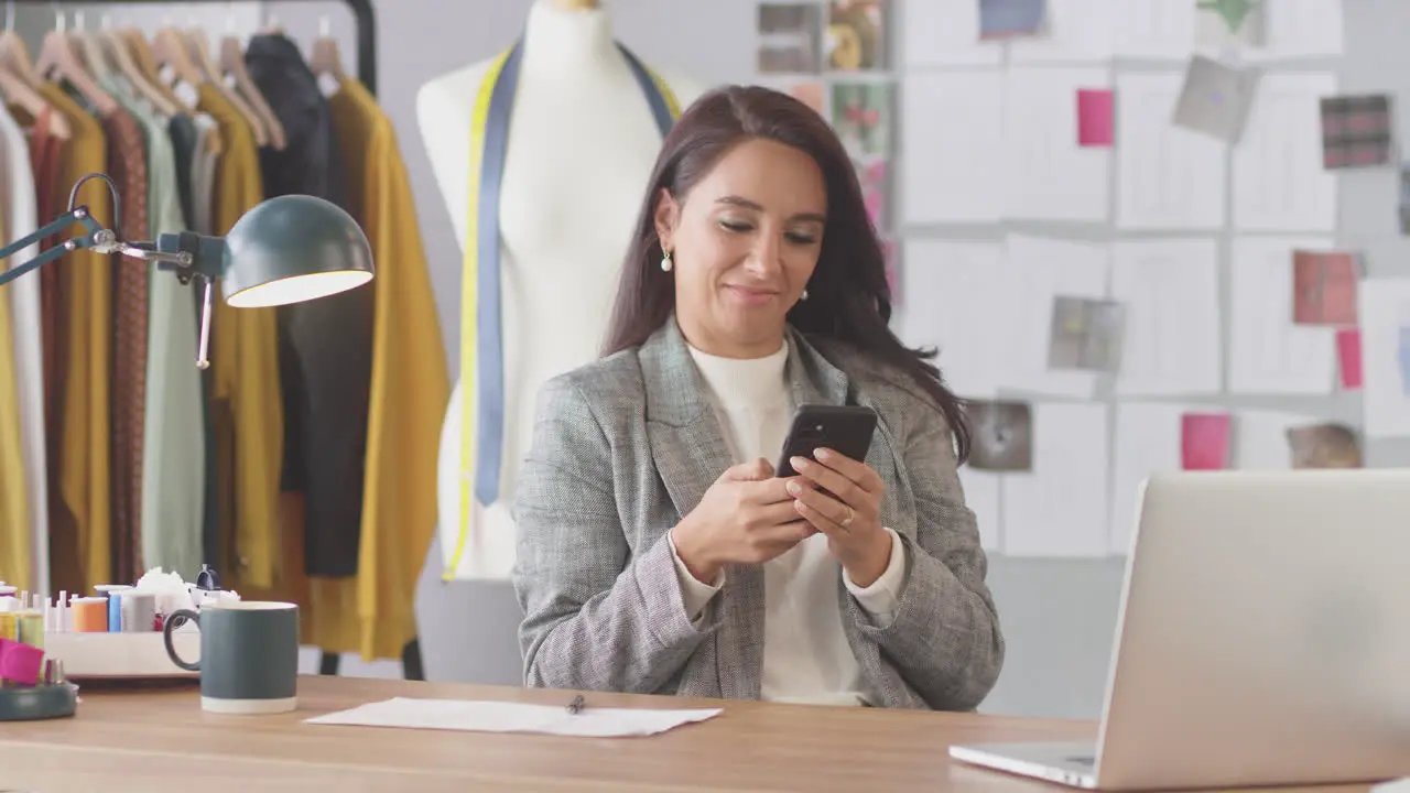 Female Fashion Designer In Studio Working At Desk On Laptop And Checking Messages On Mobile Phone