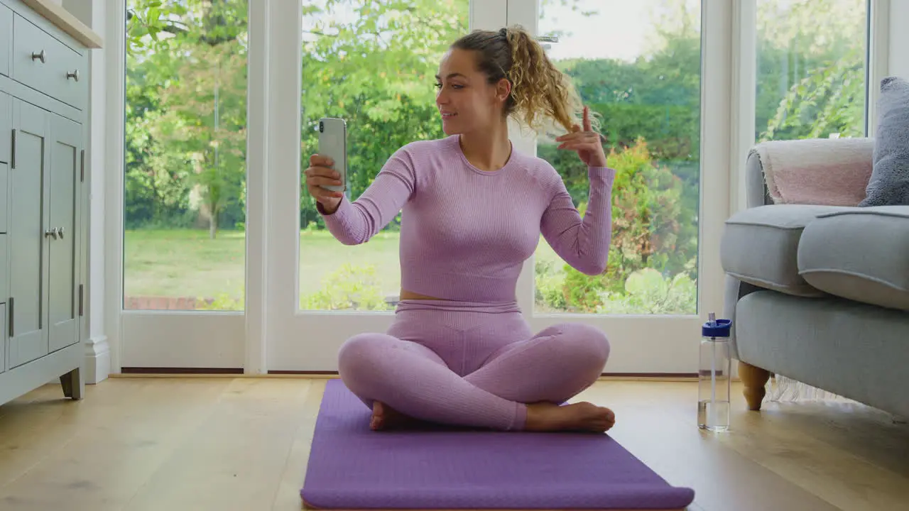 Young Woman Sitting On Yoga Mat At Home After Exercise Posing For Selfie On Mobile Phone