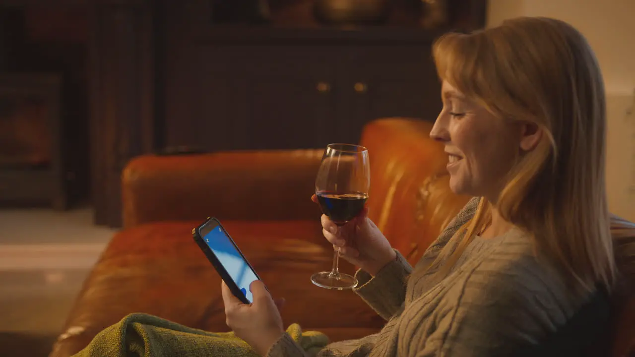 Woman At Home Sitting On Sofa With Fire With Blue Screen Mobile Phone Holding Glass Of Wine