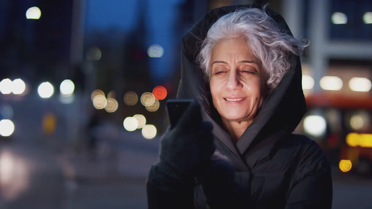 Mature Woman On City Street At Night Ordering Taxi Using Mobile Phone App