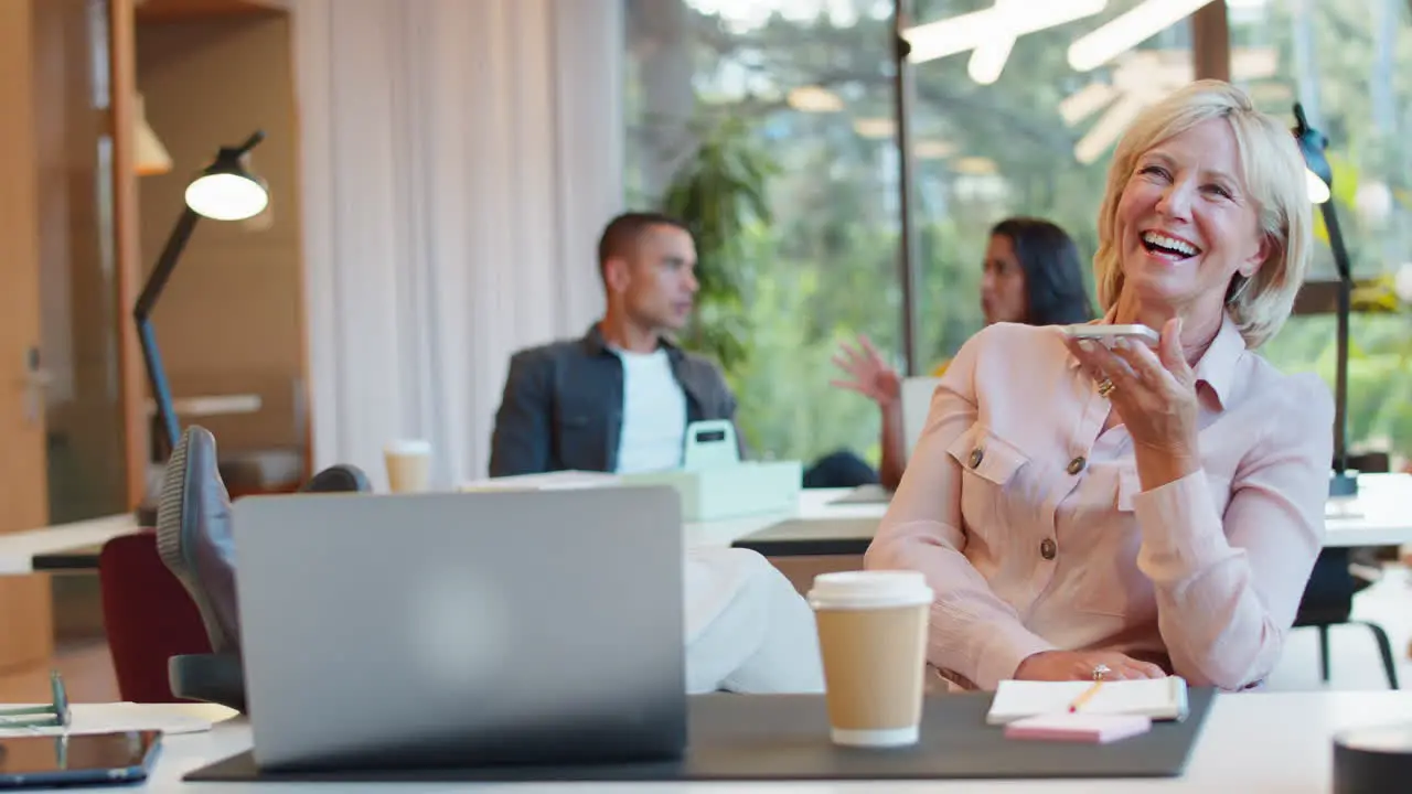 Mature Businesswoman With Feet On Desk Working In Office Talking Into Mic Of Mobile Phone