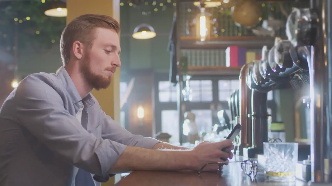 Unhappy Man Sitting At Pub Bar Drinking Alone With Glass Of Whisky Sending Message On Mobile Phone