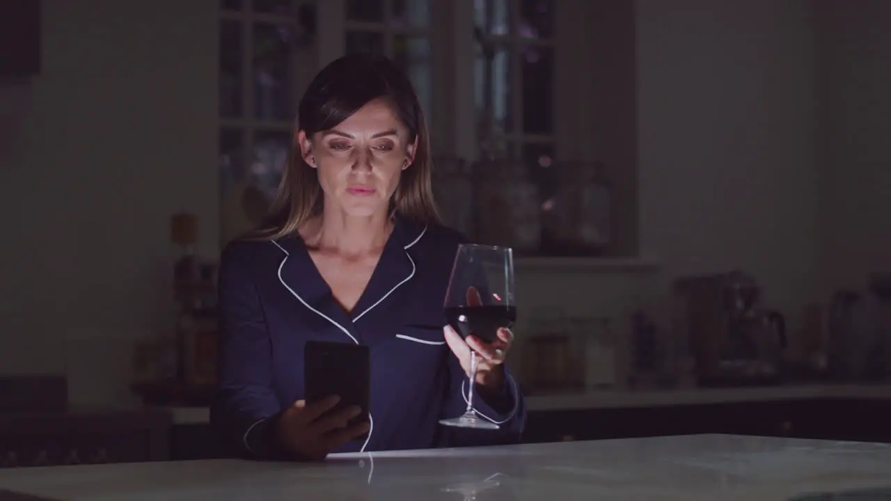 Unhappy Woman Wearing Pyjamas Sitting In Kitchen With Glass Of Wine At Night Using Mobile Phone