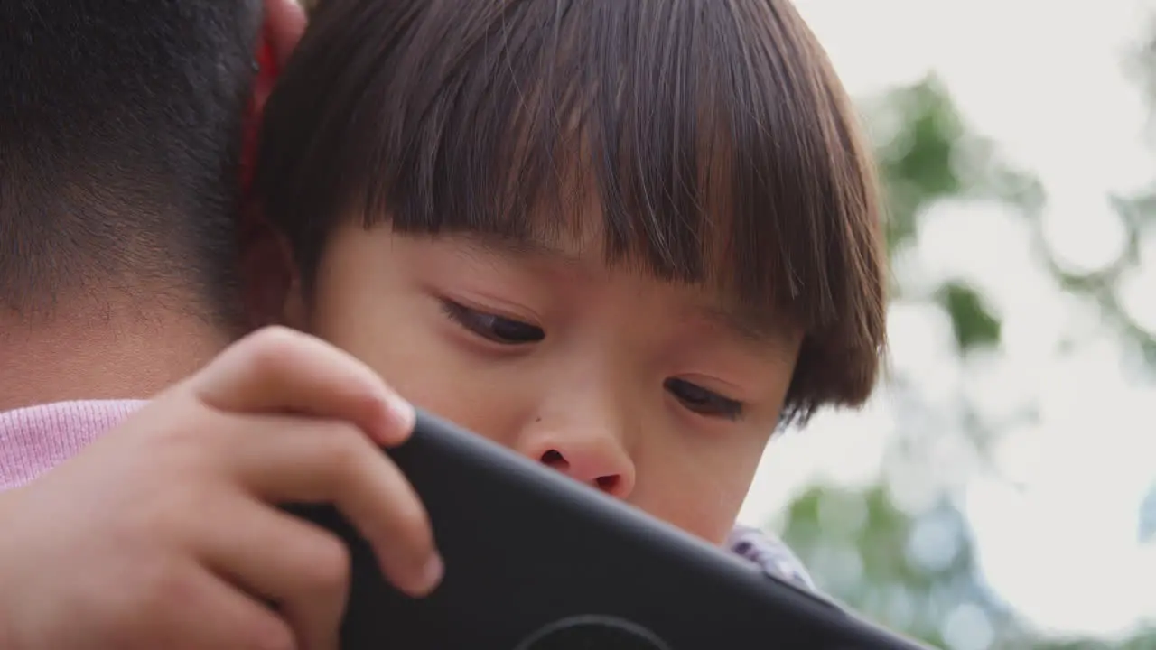 Close Up Of Asian Father Cuddling Son In Garden As Boy Looks Over His Shoulder At Mobile Phone