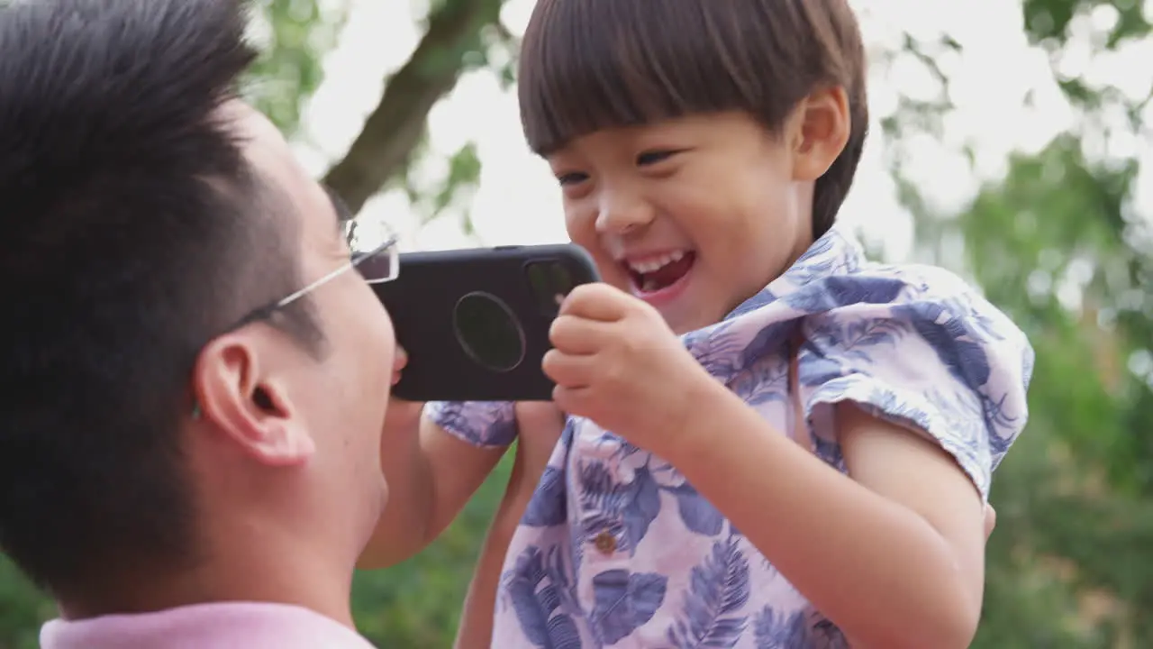 Asian Father Playing With Smiling Son In Garden As Boy Holds Mobile Phone