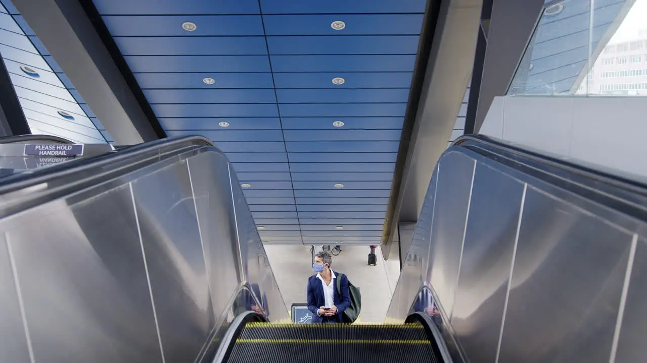 Businessman On Escalator At Railway Station With Mobile Phone Wearing PPE Face Mask During Pandemic