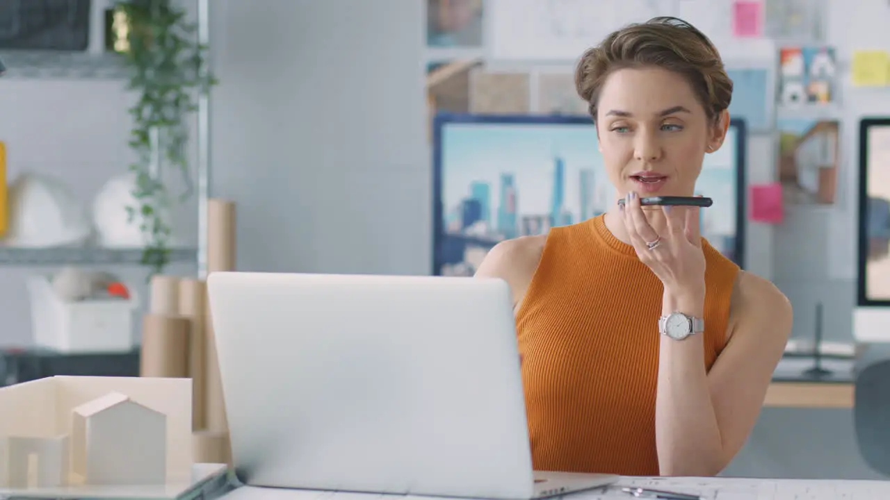 Female Architect In Office Working At Desk On Laptop Taking Speakerphone Call On Mobile Phone