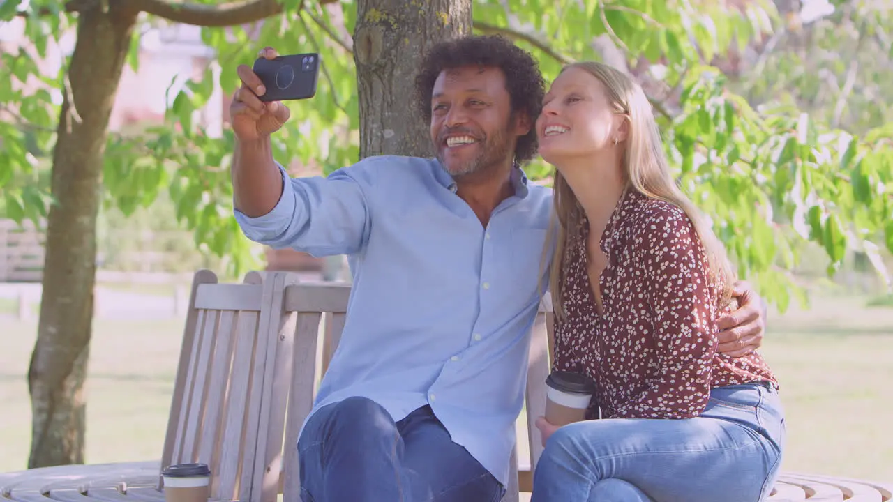 Loving Mature Couple Posing For Selfie On Mobile Phone Sitting On Seat In Park