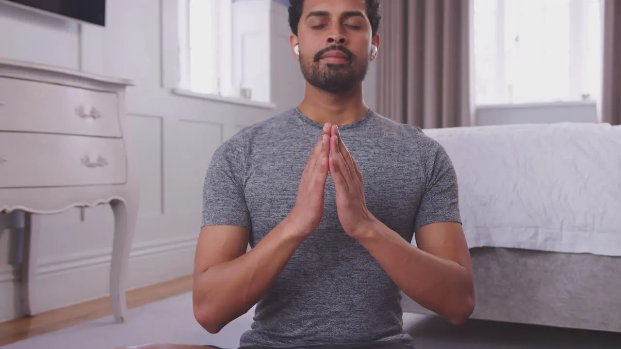 Man sitting on yoga mat in bedroom at home wearing wireless earphones and streaming from mobile phone shot in slow motion