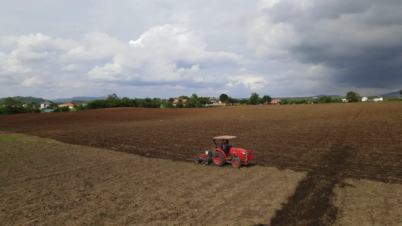 Static shot of a red tractor tilling a rich fertile soil moving diagonally leaving furrows contrasting between old and new plowed land in rural countryside in Thailand Southeast Asia