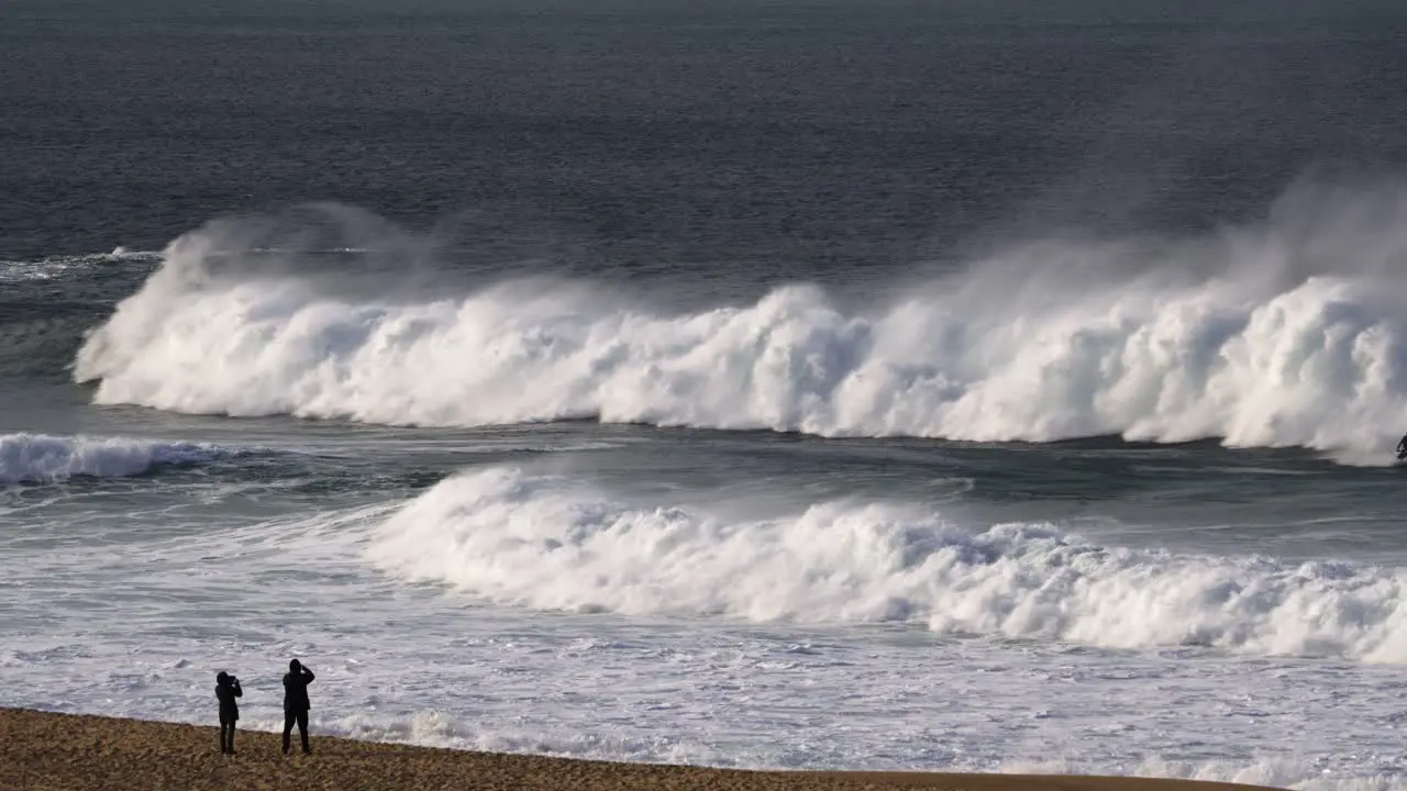 Two people on a sandy beach watching big rising sea waves