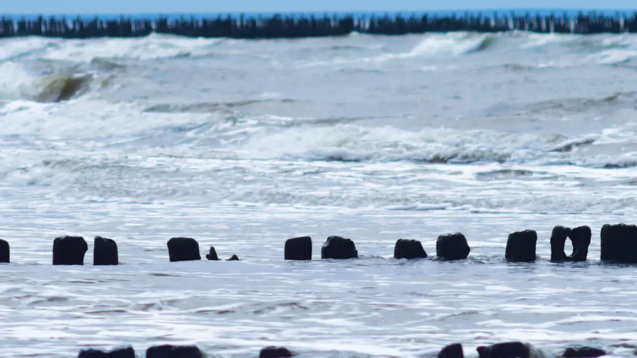 Stormy waves breaking against old wooden pier on the beach overcast spring day Baltic sea Latvia Pape medium shot from a distance