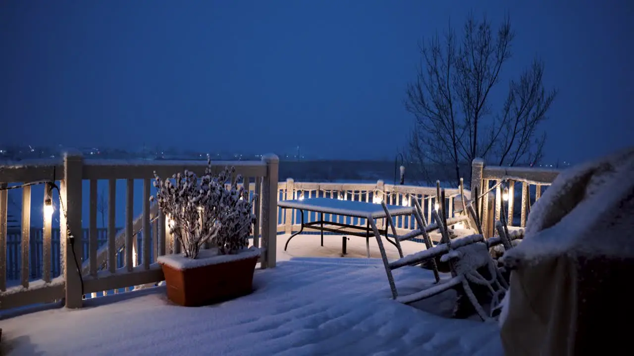 A backyard deck on a snowy night with the porch light son and the dining table covered in snow static wide angle view