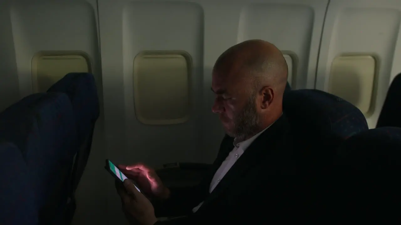 A man working and browsing on a smartphone on a passenger airliner plane at night in a dark cabin