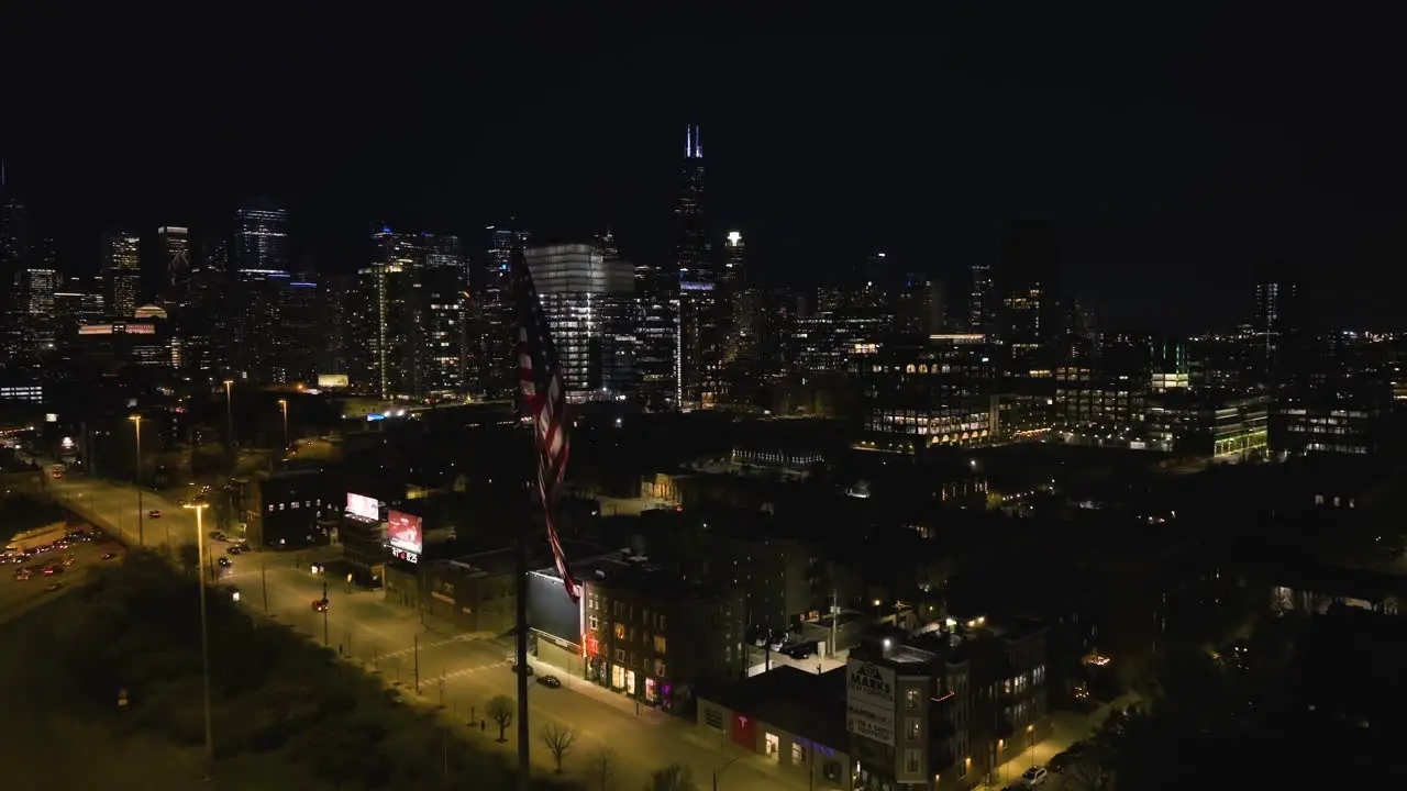 Aerial view flying past a waving flag of USA approaching the night lit downtown of Chicago