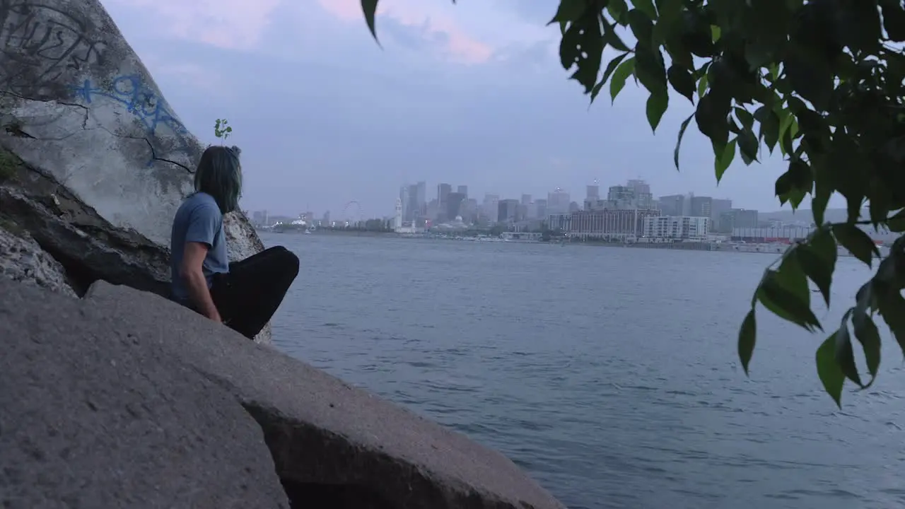 A Man in a Blue Shirt Sitting on Some Rocks Across the River from Downtown Montreal on a Cloudy Foggy Evening