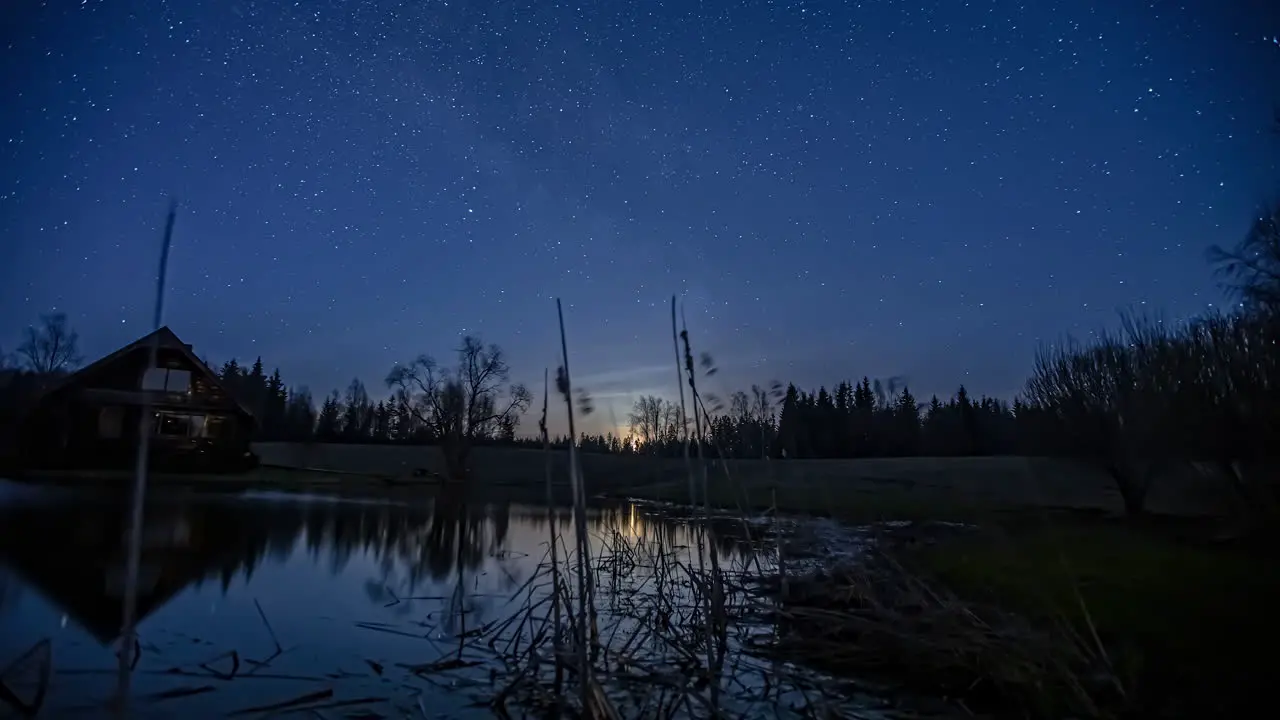 Milky Way and moon moving across sky over cabin by a lake at night