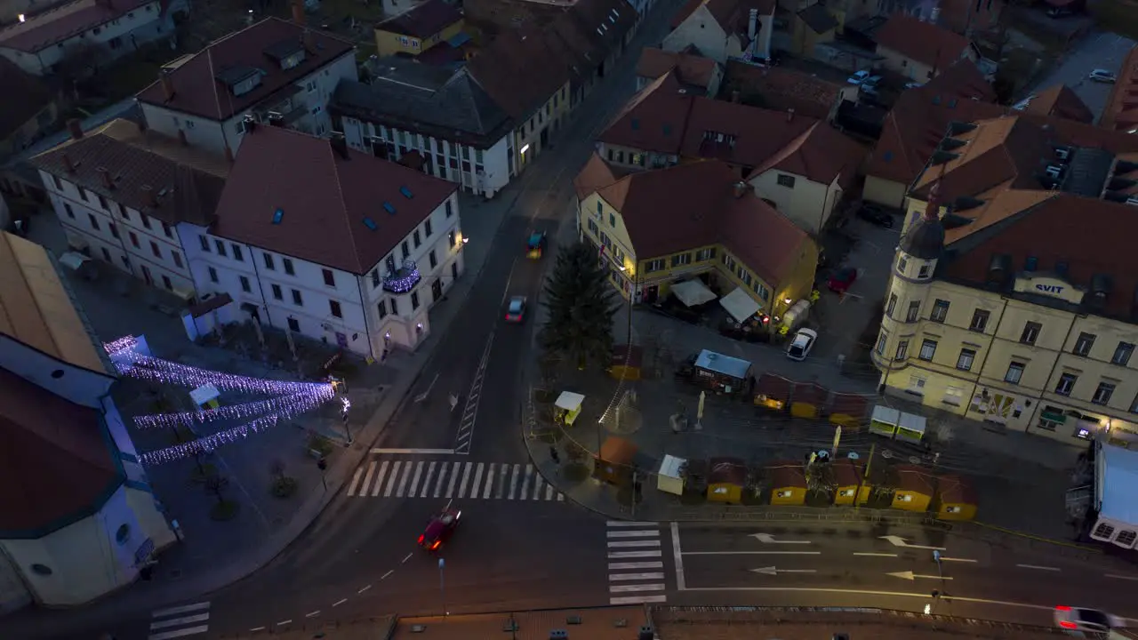 Christmas lights ignite on small town square dusk to night aerial timelapse of Xmas market cars pass by on street steady shot