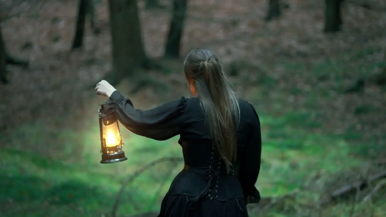 Girl with long hair and a black dress walks through a dark forest with a lamp in her hand