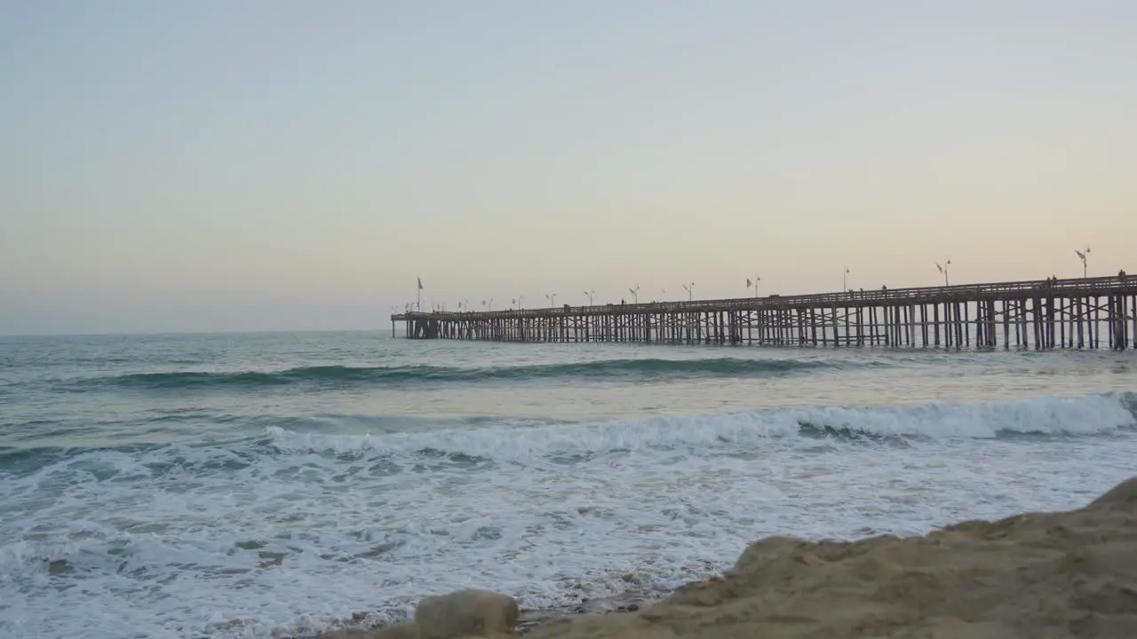 slow motion Panning shot of waves rolling through the Pacific Ocean crashing into the shores of Ventura Beach with the pier and sunset in the background located in Southern California