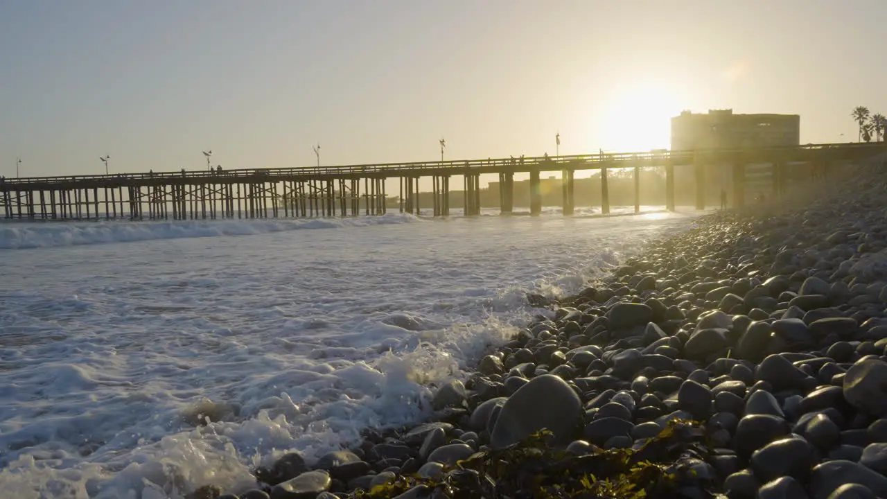 Dolly shot moving forward along the rocky shore of a Southern California Beach while waves are crashing in with Venture Pier in the background located in Southern California