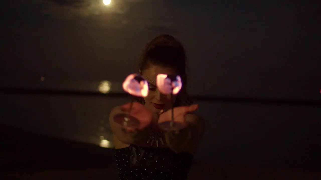 Woman Twirling Fire Palm Torches Against Full Moon lit Lake Night Exterior Medium Shot Slowmo