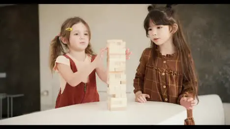 Two preteen girls friends children playing board game Jenga on table in kitchen at home slow motion