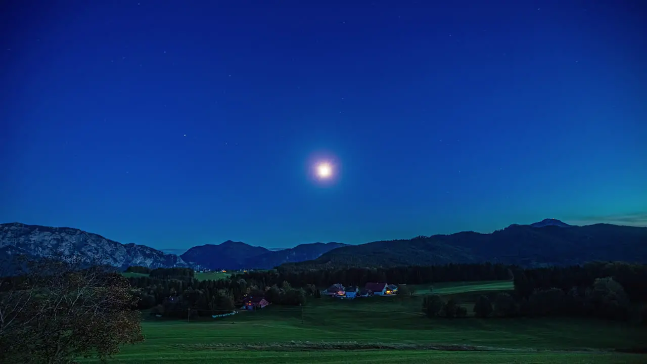 Static wide shot of a picturesque landscape in austria at night with intense blue sky at full moon over the majestic mountain ranges and dense forest with meadows