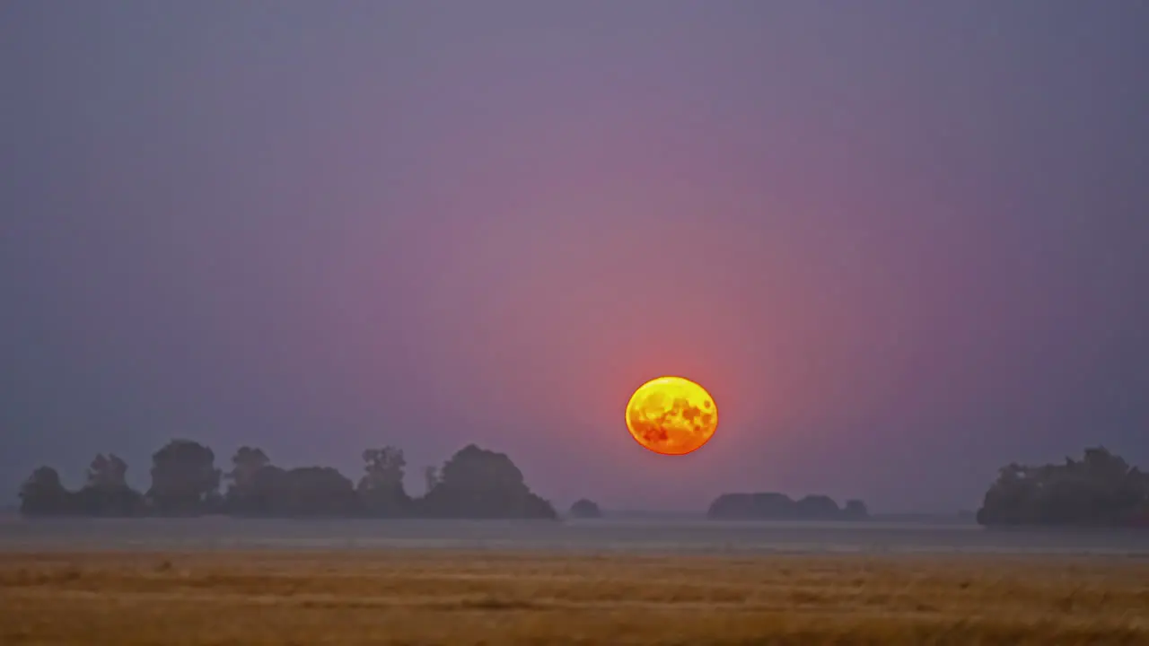 Super moon rises over a farm field with a low layer of fog or mist to glow over the countryside time lapse