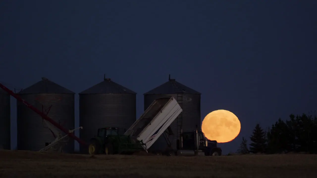 Telephoto timelapse of full moon rising behind grain silos during harvest