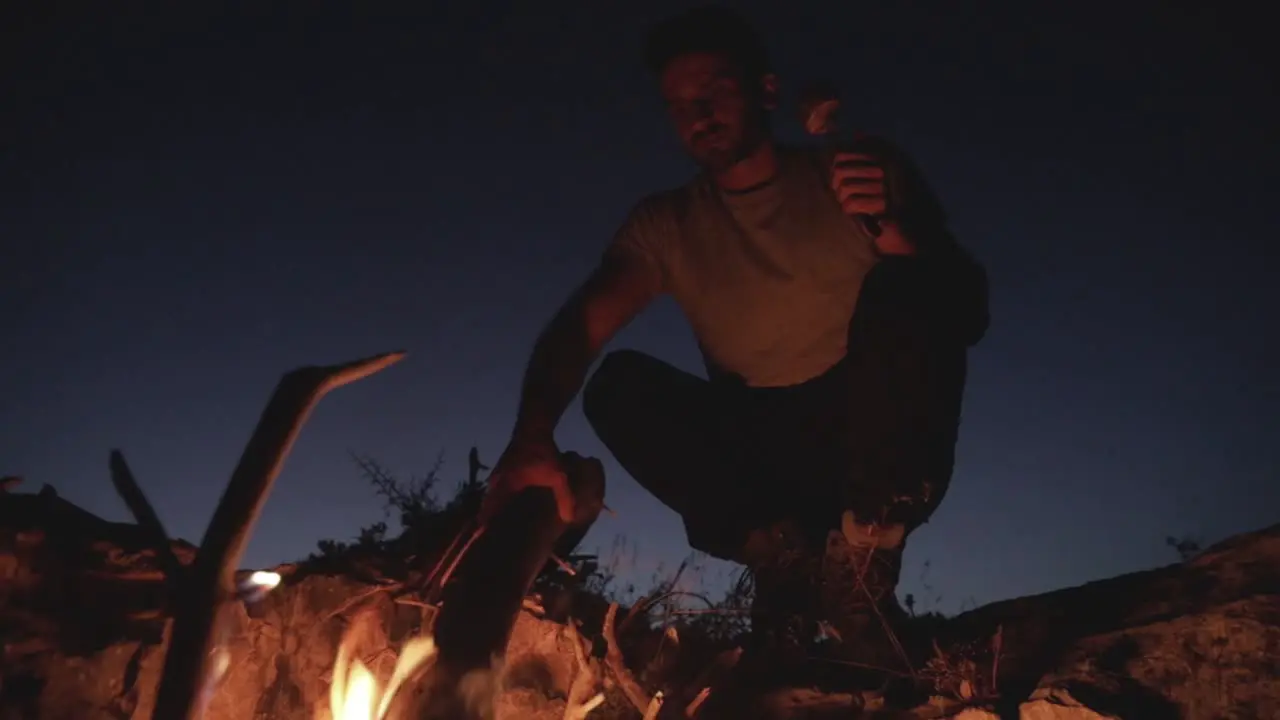 Caucasian man moving dead wood log in firepit on bright clear evening sky with half moon in distance low angle close up pan