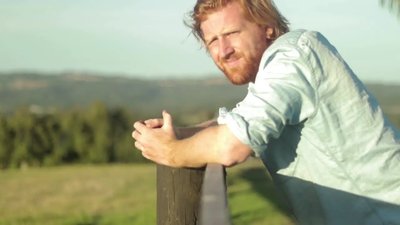 A young farmer in a blue shirt rests on a wooden fence post and looks out across the country in slow motion