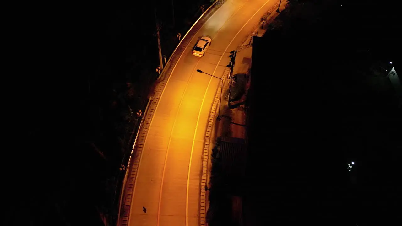 Aerial drone shot of a white car driving down an empty road at night in a rural area with yellow street lights