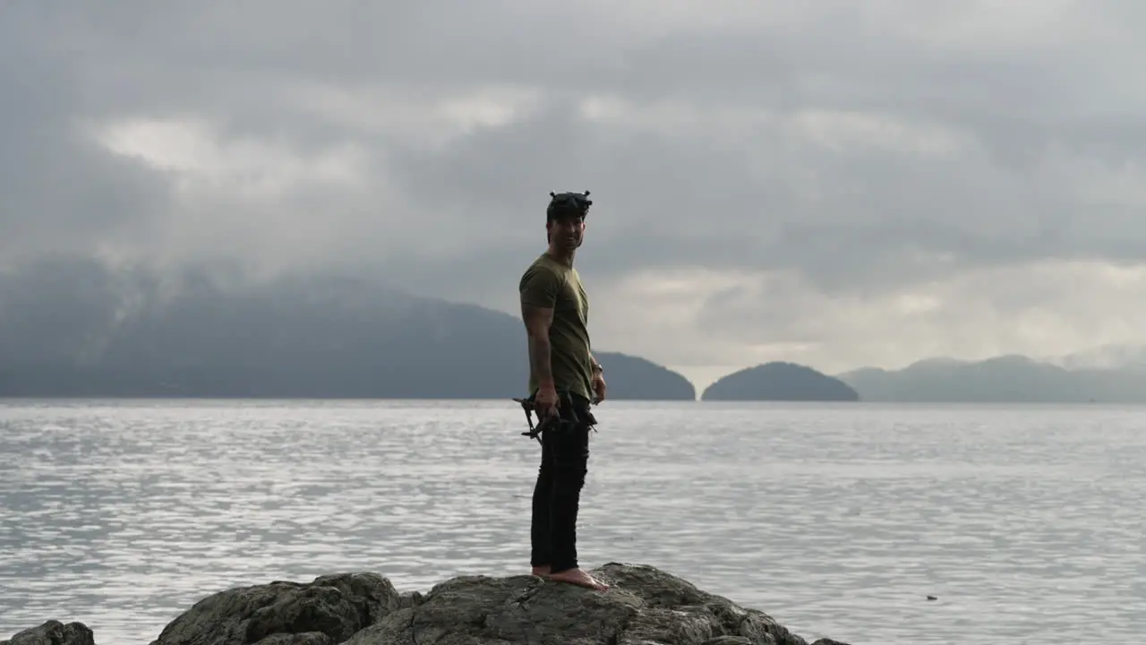 Caucasian Man With FPV Quadcopter Standing On Rock Overlooking Calm Sea With Overcast