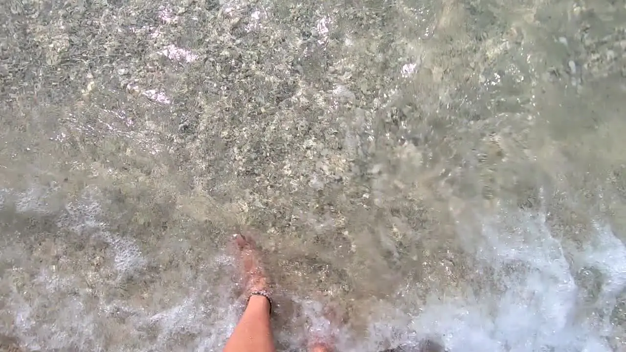 Slow-motion view of two feet standing on a sandy tropical beach while waves of clear warm water splash onto the shore