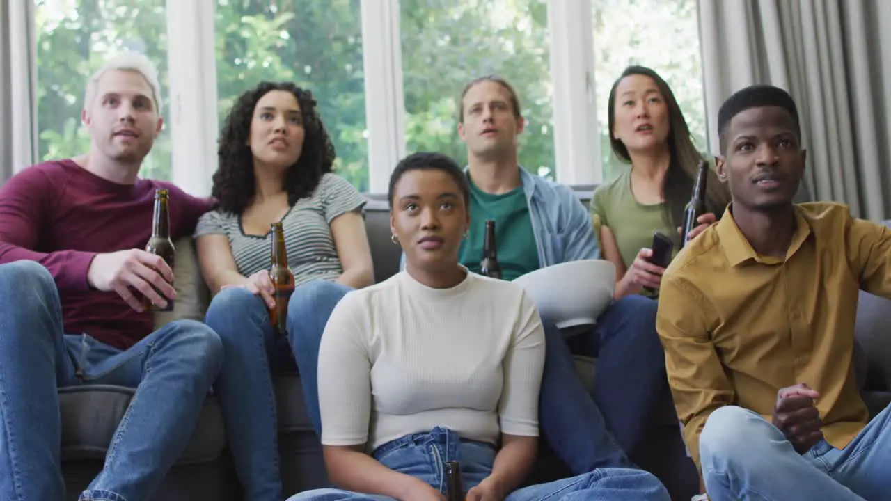 Group of diverse young people holding beers cheering while watch tv at home