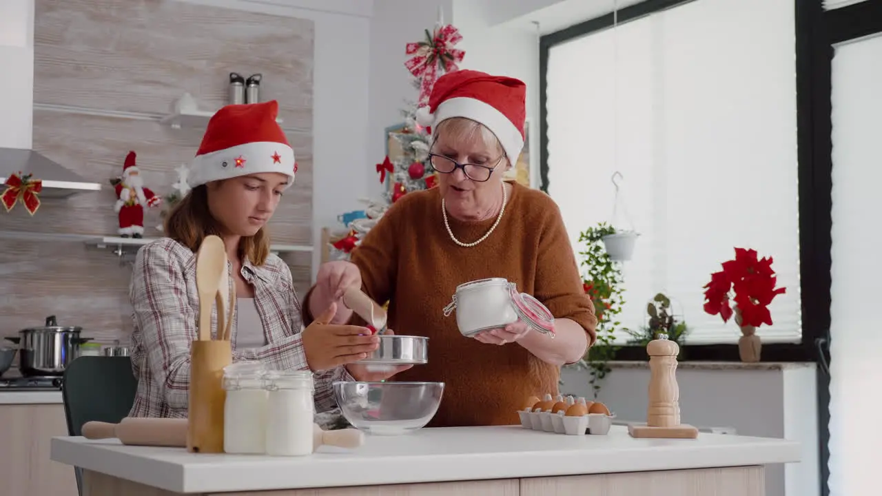 Granddaughter using kitchen strainer while grandmother putting flour ingredient