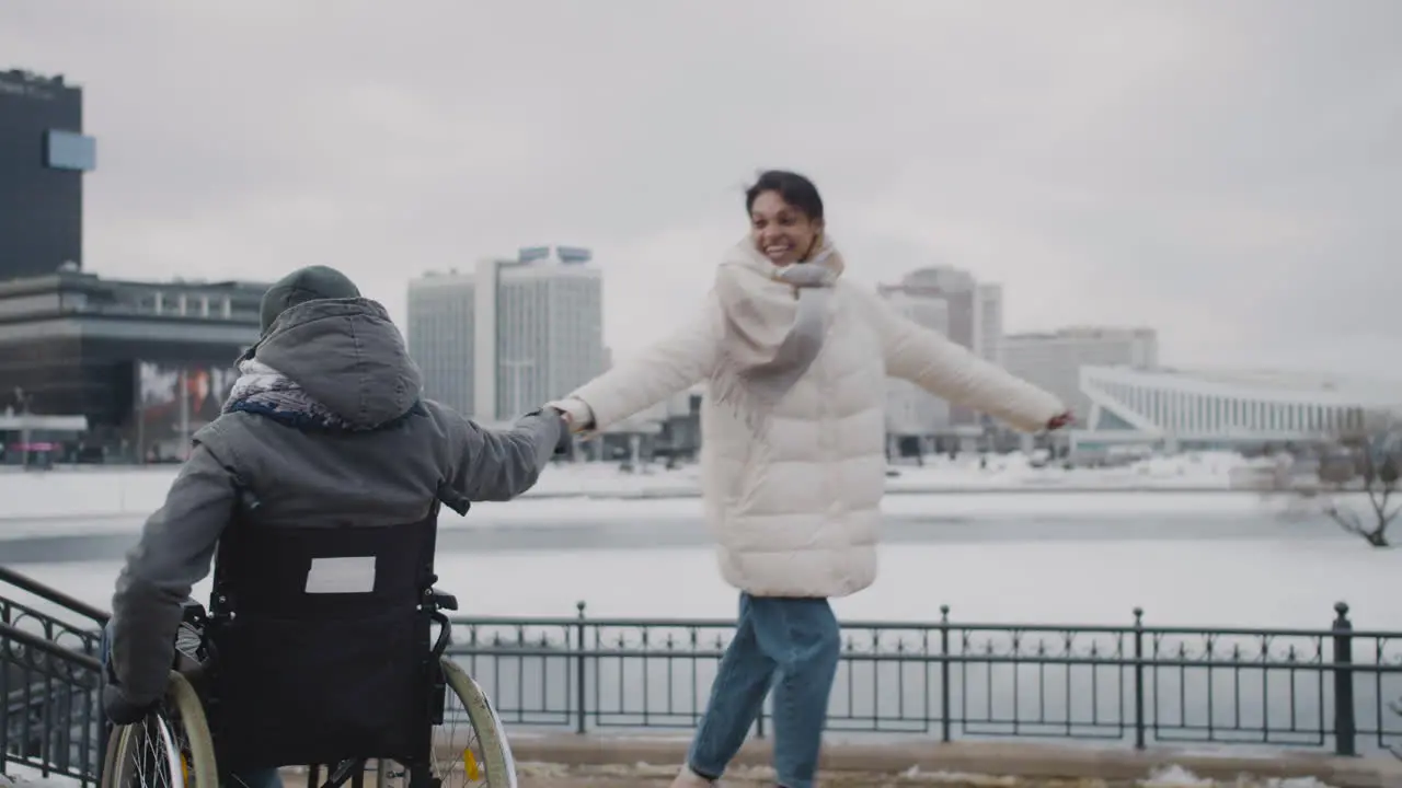 Happy Muslim Woman And Her Disable Friend In Wheelchair Spinning Holding Hands In City In Winter 1