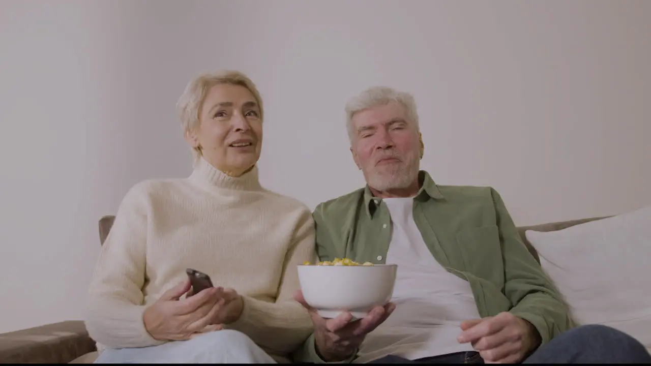 Happy Senior Couple Eating Caramel Popcorn And Watching Tv While Sitting On Sofa At Home 1