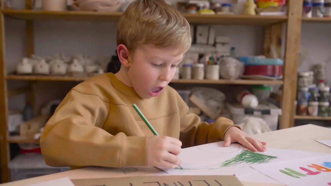 Blond Kid Drawing With Green Pencil Sitting At A Table In A Craft Workshop Where Are Signs With Environmental Quotes