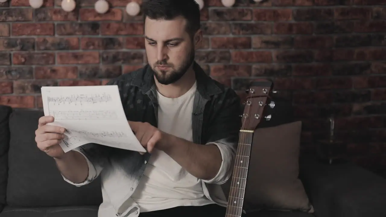 Young Musician Man Looking At Sheet Music To Play Guitar At Home