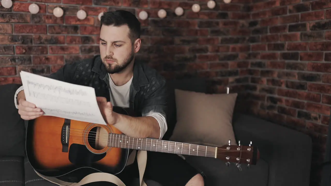 Young Musician Man Looking At Sheet Music And Playing Guitar At Home