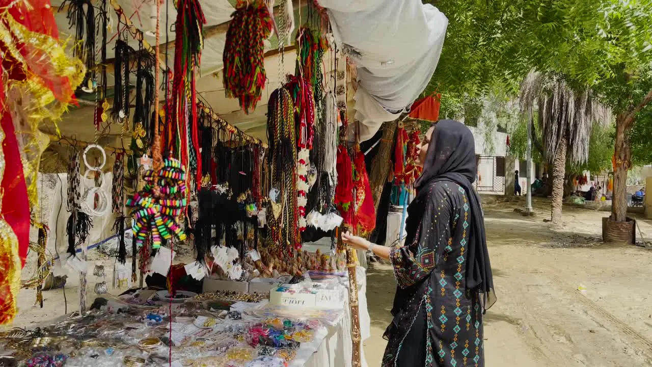 Pakistani Woman Wearing Head Scarf Walking Towards Market Stall Selling Colourful Beads In Hingol Balochistan