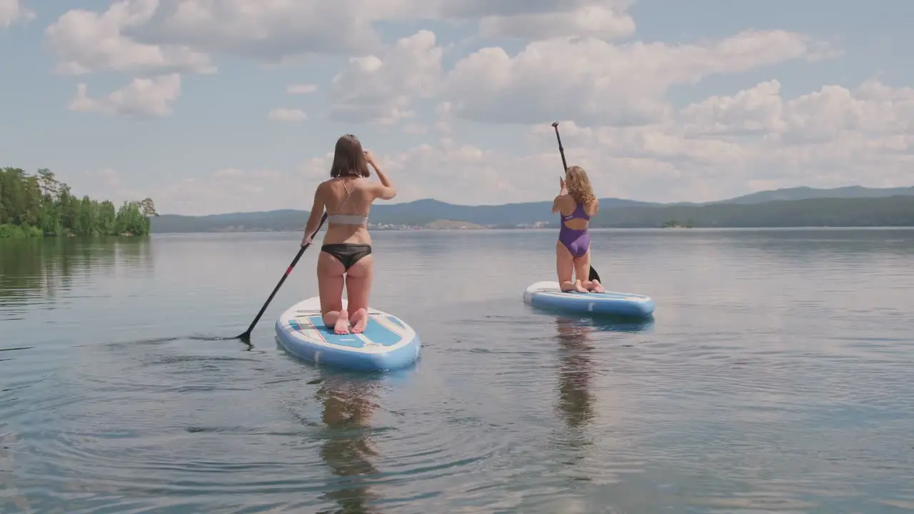 Rear View Of Two Girls In Swimsuit Doing Paddle Surfing On Knees In The Sea