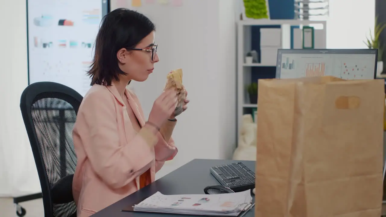 Businesswoman eating bite of tasty sandwich drinking coffee in front of monitor