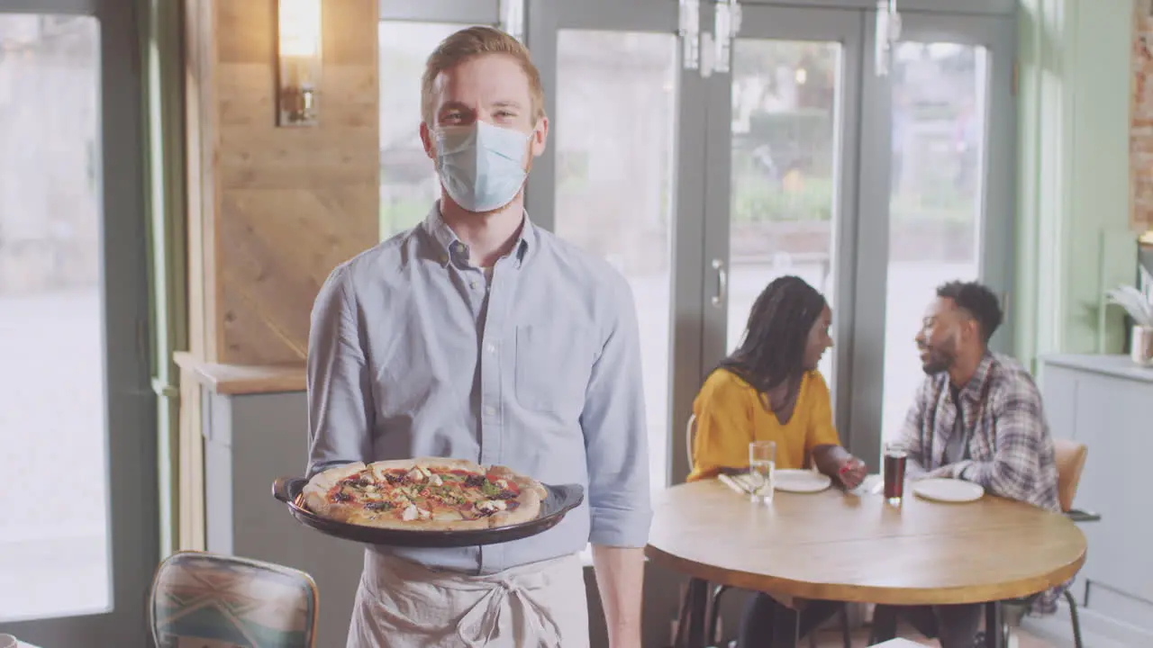 Portrait Of Waiter In Restaurant Wearing Mask Serving Pizza To Couple During Health Pandemic