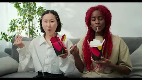 Diverse female friends eating noodles together at home using chopsticks