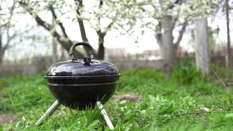 A metal barbecue grill stands in the backyard on the ground with grass by the bushes