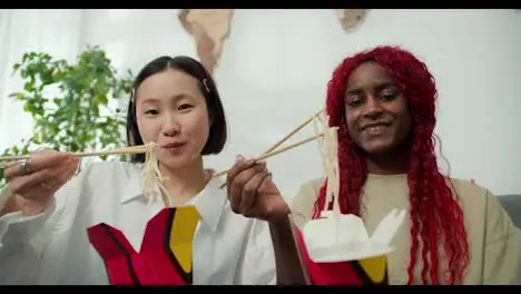 Diverse female friends eating noodles together at home using chopsticks positive mood